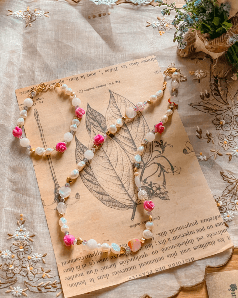 Een handgemaakte ketting en armband met kleurrijke kralen en rozenapplicaties, rustend op een vintage boek en decoratieve stof.